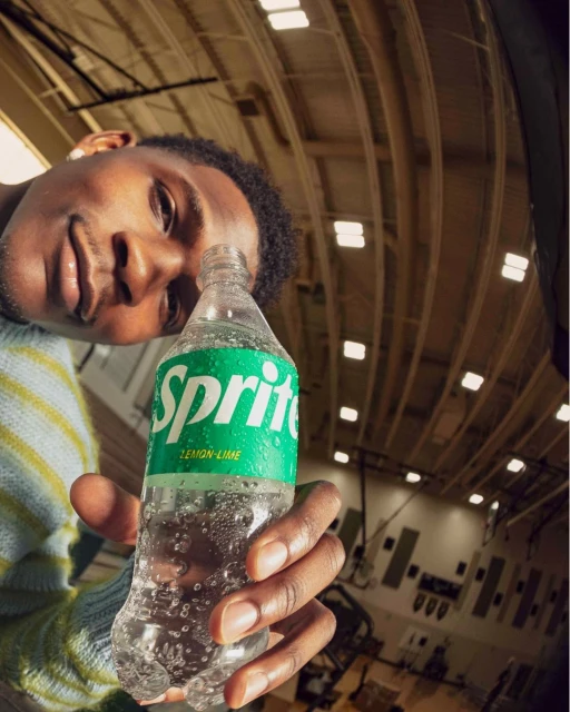 Man holding a Sprite bottle in an indoor sports arena. The fisheye-lens image focuses on the bottle's label and cap, with the man's face.