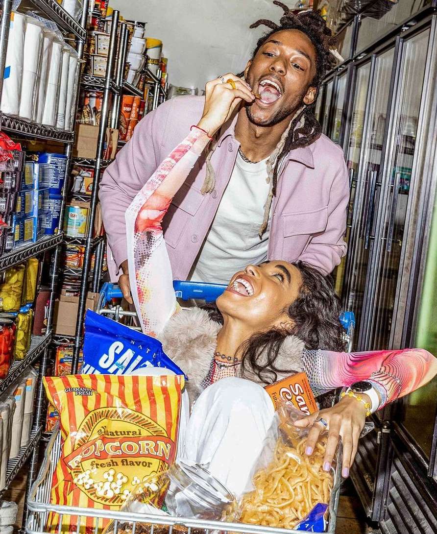 Woman in shopping cart filled with snack foods laughs while man eats a chip in a brightly lit grocery store aisle.
