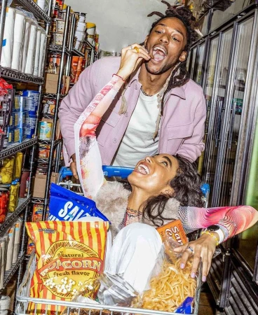 Woman in shopping cart filled with snack foods laughs while man eats a chip in a brightly lit grocery store aisle.