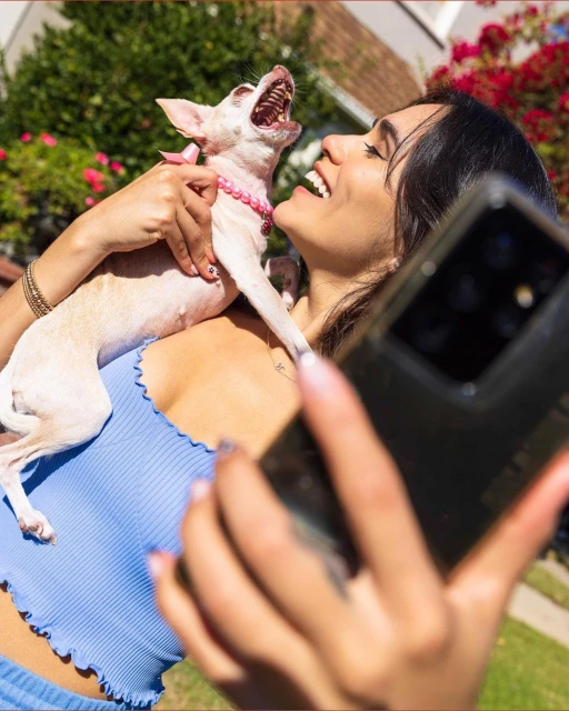 A smiling woman in a blue top holds a small dog outdoors, taking a selfie with her smartphone in a sunny backyard setting.