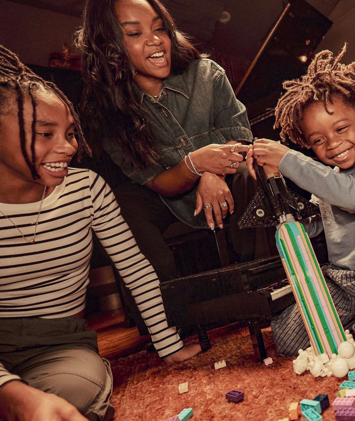 Smiling mother and two children playing with toy rocket and blocks on a brown carpet in an indoor setting with a piano in the background.