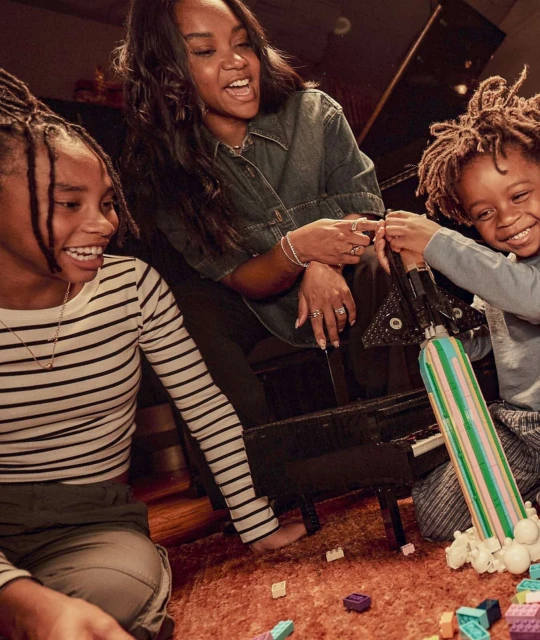 Smiling mother and two children playing with toy rocket and blocks on a brown carpet in an indoor setting with a piano in the background.