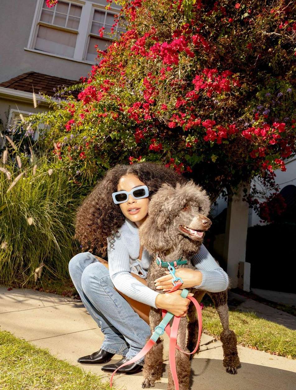 Woman with curly hair embraces a poodle on a sidewalk in front of a house with red flowers on the bush alongside the building.