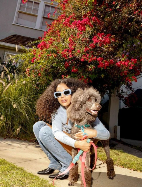 Woman with curly hair embraces a poodle on a sidewalk in front of a house with red flowers on the bush alongside the building.
