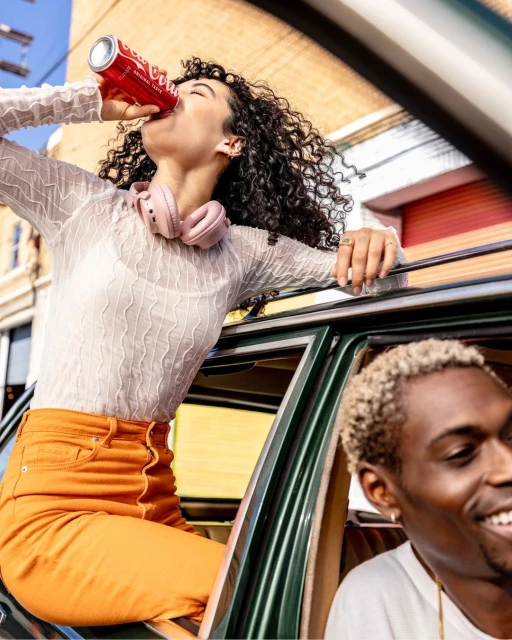 Young woman drinks from a red can sitting on a car door as a young man smiles in the background. Bright outdoor daylight.