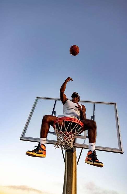 Low angle shot of a man in a white tank top sitting on a basketball rim against a clear sky