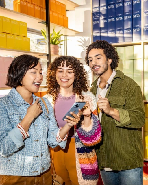 Three friends smile while looking at a smartphone in a well-lit shop with shelves displaying products, creating a friendly and engaging scene.