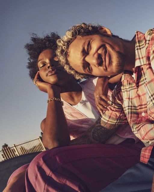 Portrait of a couple embracing under a blue sky. The man wears a red and white plaid shirt and has light colored hair. The woman has dark hair.