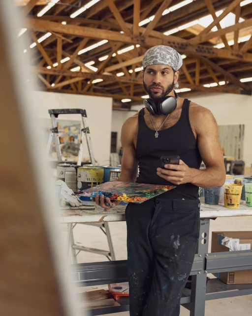 Male artist wearing headphones and bandana holds a paint palette and cell phone in an art studio filled with paint cans and wooden beams.