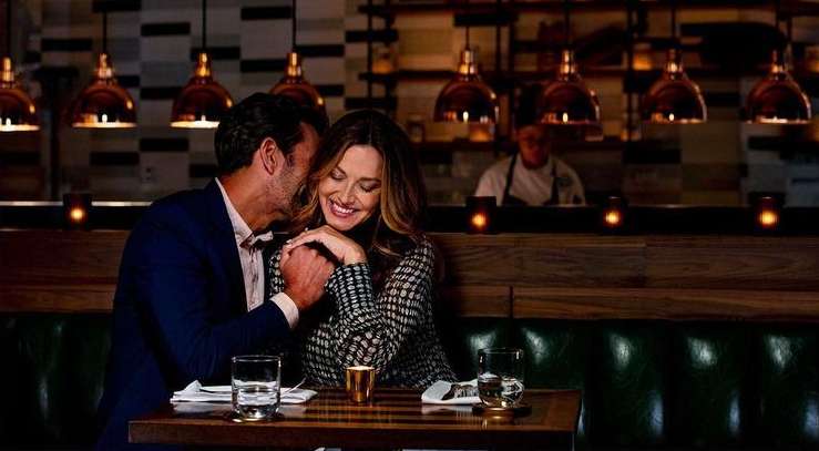 Man in a suit whispering into the ear of a woman in a patterned blouse at a restaurant with mood lighting.