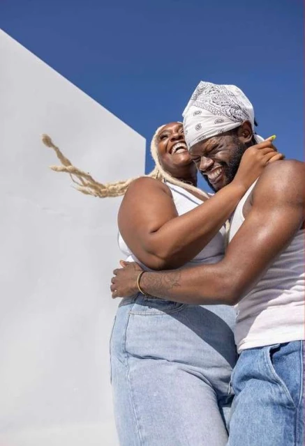 A happy couple embraces against a bright white wall under a clear blue sky. They are laughing, wearing casual clothing, and appear joyful.