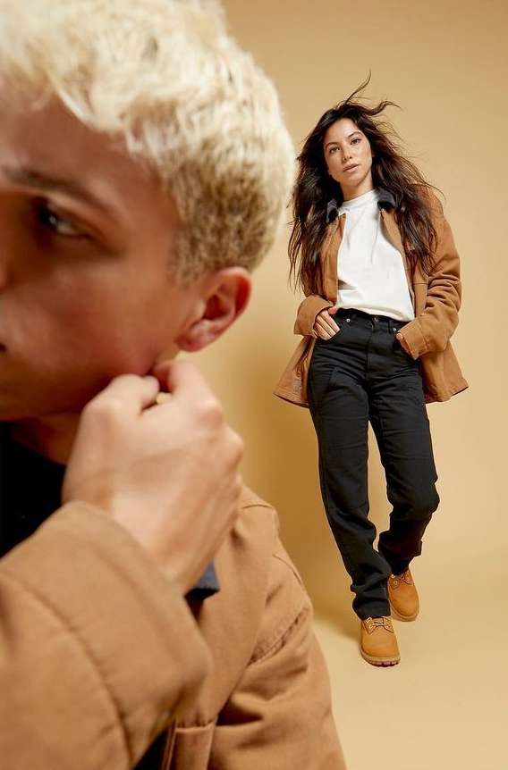 Studio shot of a woman in casual wear with a man in a brown jacket slightly out of focus in the foreground.