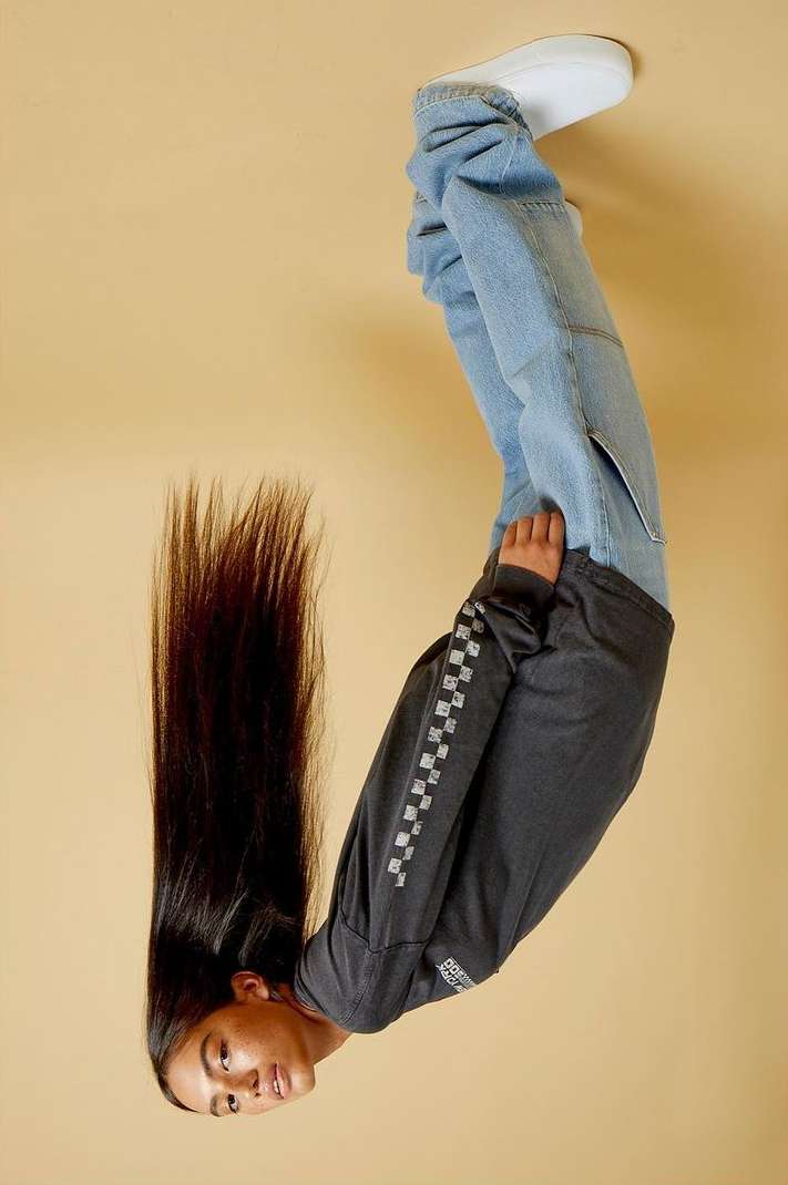 Young person poses upside down in a studio wearing a long-sleeved shirt, jeans, and shoes against a tan backdrop with long dark hair.