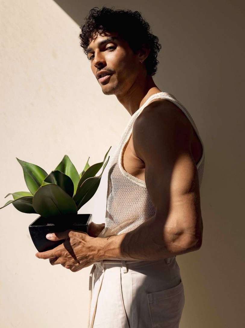 Adult male poses in a mesh tank top, holding a snake plant in a black pot against a light beige wall with soft, diffused lighting.