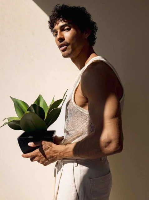 Adult male poses in a mesh tank top, holding a snake plant in a black pot against a light beige wall with soft, diffused lighting.