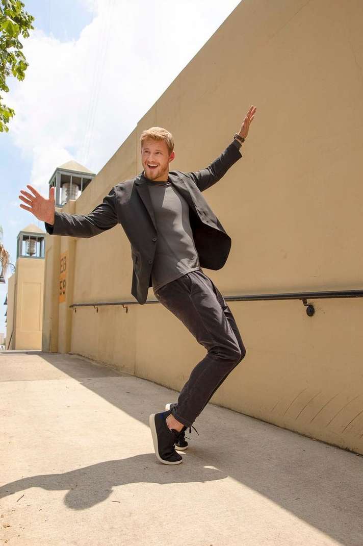 Man in suit jacket and casual pants strikes a playful pose outside against a neutral wall, expressive posture and bright sunlight.