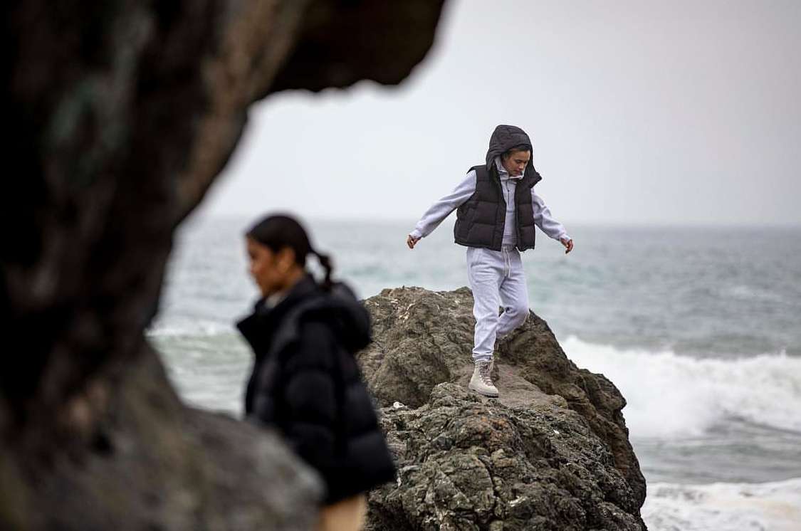 Two young women in outerwear stand on rocks at the ocean's edge on an overcast day, with waves and a gray sky in the background.