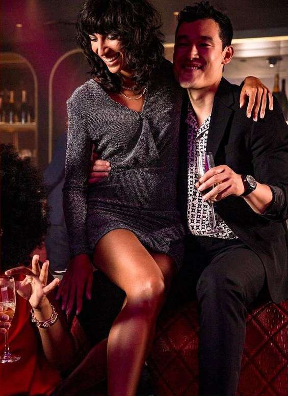 Smiling woman in a sparkly dress sits on a red quilted surface with an arm around a man in a black suit holding a champagne flute indoors.