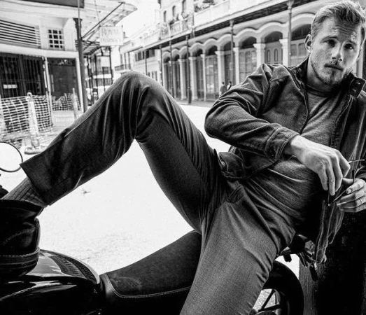Black and white portrait of adult male with stubble, sunglasses in hand, sitting on a motorcycle on a city street with storefronts in background.