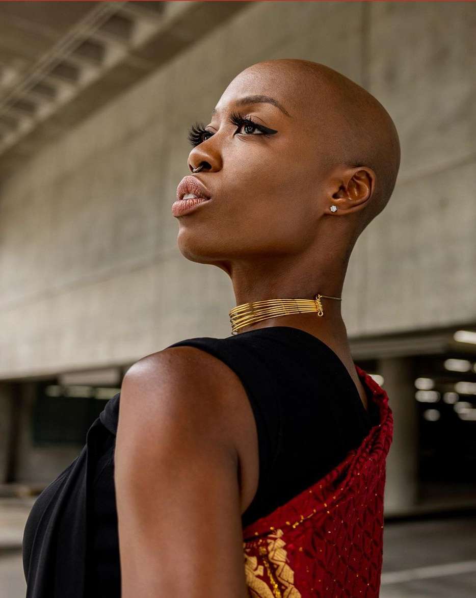 Elegant portrait of a bald African woman in gold jewelry, wearing a black top and red shawl, against a concrete background.