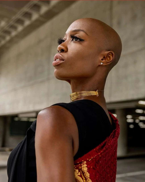 Elegant portrait of a bald African woman in gold jewelry, wearing a black top and red shawl, against a concrete background.