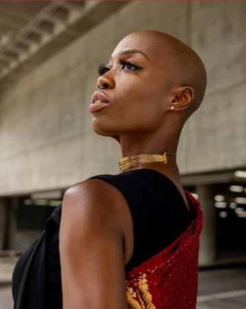 Elegant portrait of a bald African woman in gold jewelry, wearing a black top and red shawl, against a concrete background.