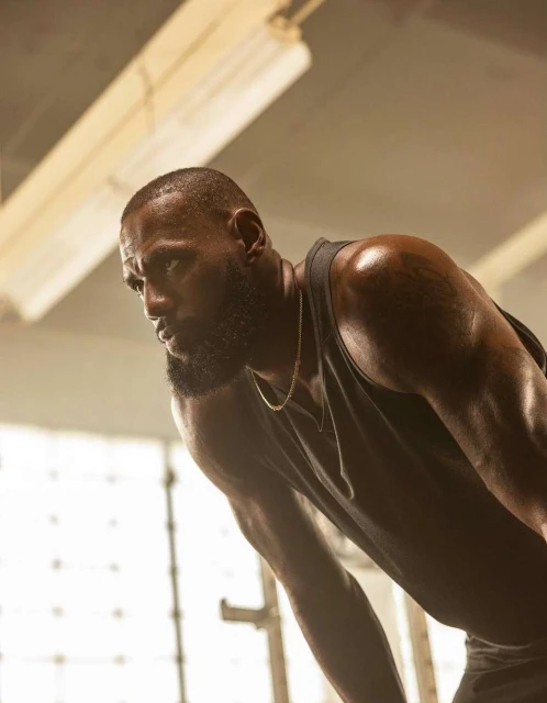 Athletic African-American man with muscular build, wearing a tank top and chain, bending over in an indoor gym setting.
