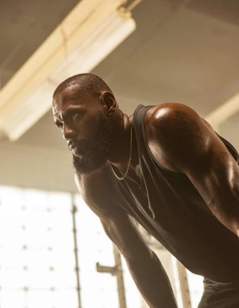 Athletic African-American man with muscular build, wearing a tank top and chain, bending over in an indoor gym setting.