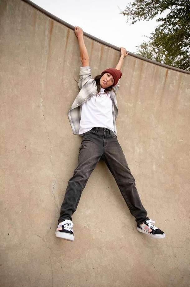 Young woman in casual wear hangs upside down from the top of a skateboard ramp, showcasing her outfit, including a beanie and sneakers.
