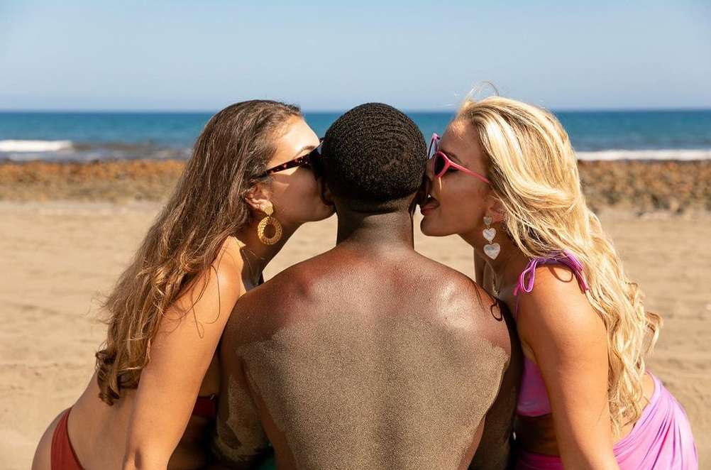 Two women kiss a man on the cheeks on a sunny beach near the ocean. The women wear swimwear and the man's back is visible.