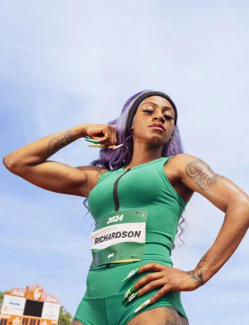 A muscular female athlete with long purple hair, wearing a green athletic outfit, stands on a track under a bright, sunny sky.