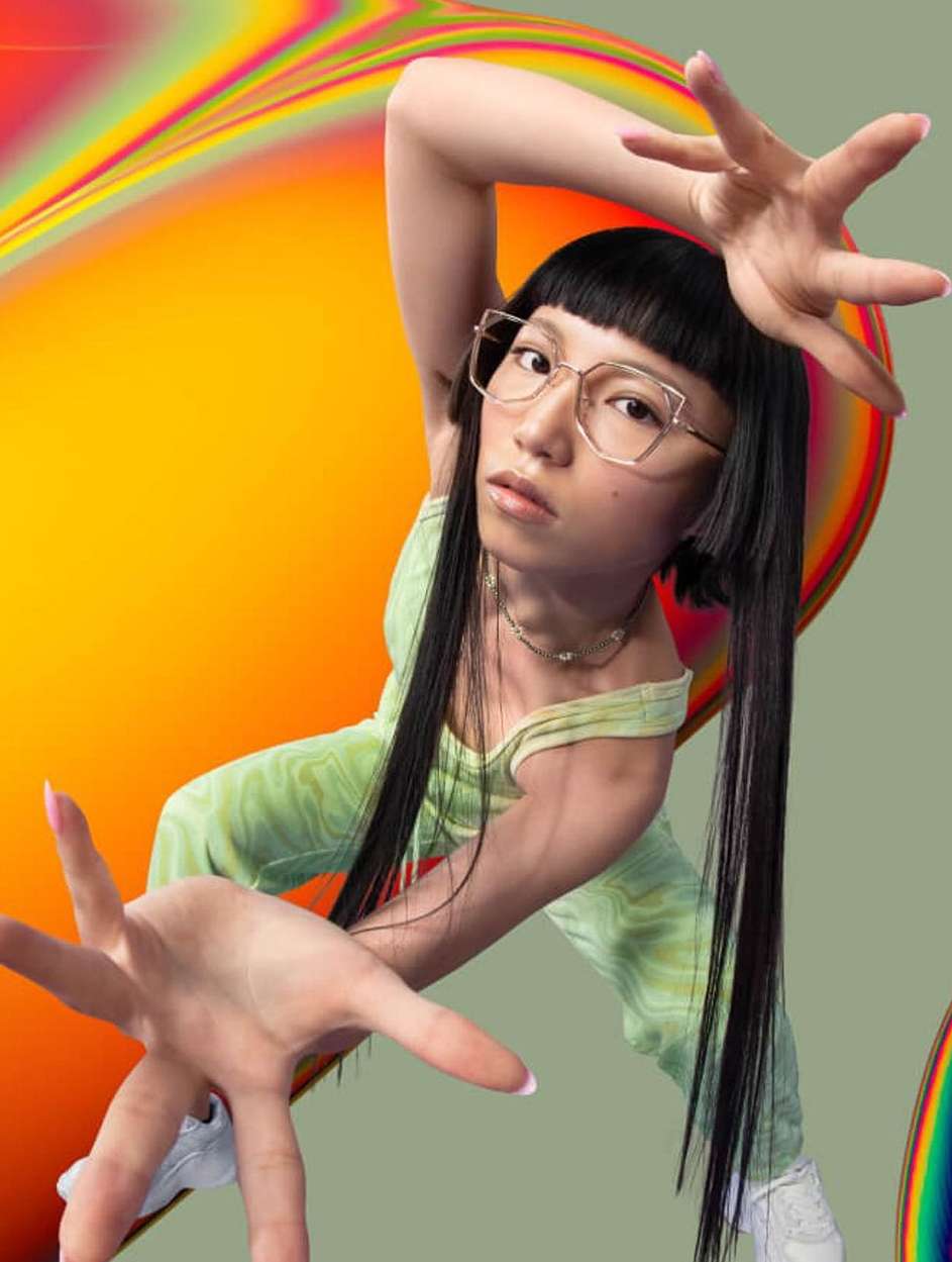 Young woman with dark hair and glasses poses with hands outstretched against a colorful abstract background in a studio shot.