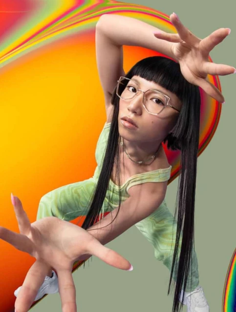 Young woman with dark hair and glasses poses with hands outstretched against a colorful abstract background in a studio shot.