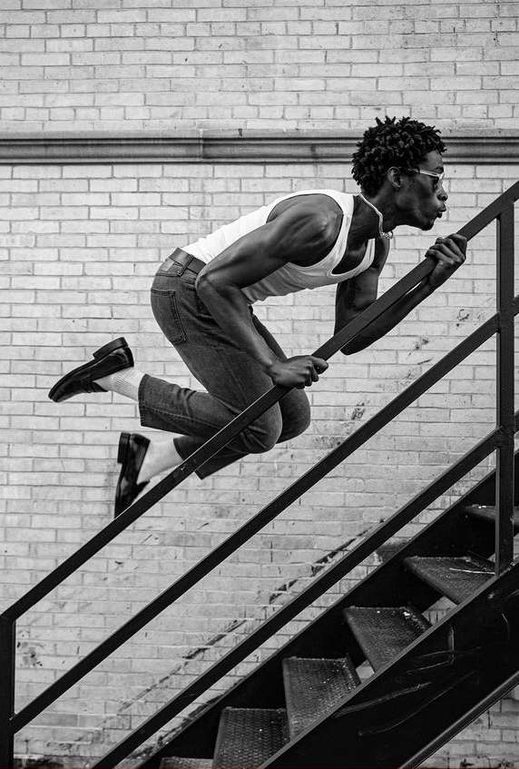 A man in a white tank top and blue jeans jumps on black stairs in a black-and-white shot, with a brick wall in the background.