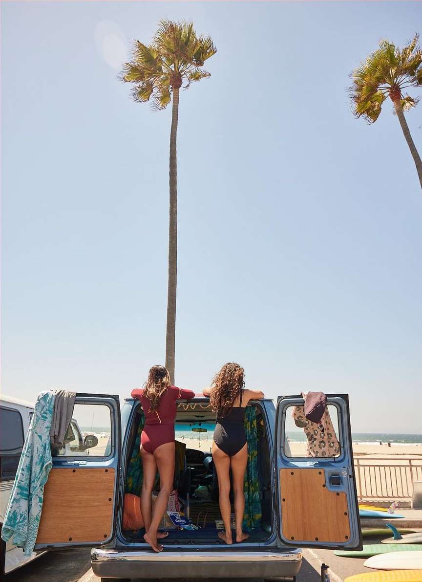 Two women in swimsuits lean against a van at the beach with surfboards nearby. Palm trees and a clear blue sky in the background.