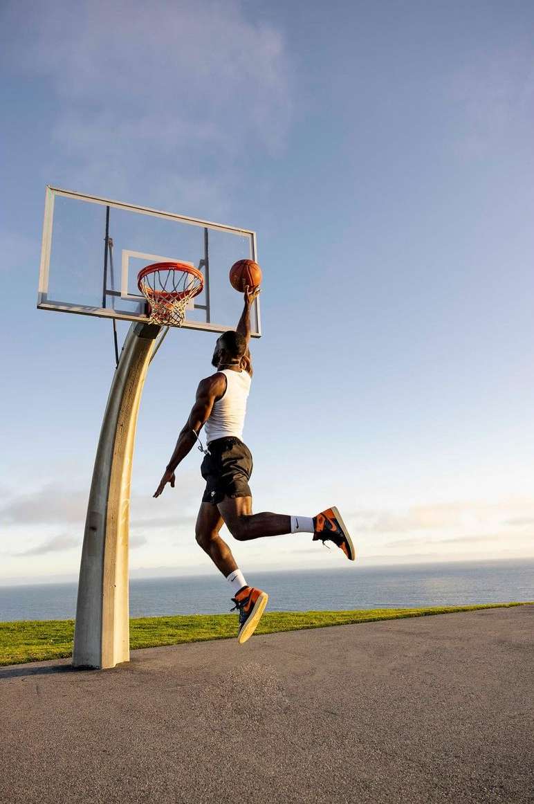 Athletic man in mid-air performing basketball dunk near the ocean on an outdoor court in white tank top, black shorts, and orange sneakers.