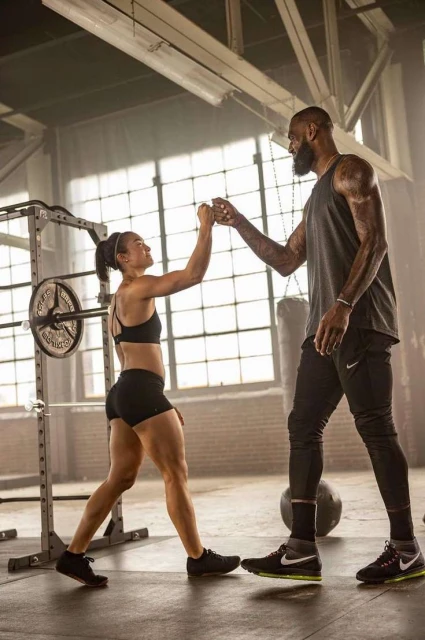 LeBron James and a female athlete fist-bumping in a gym. They are both wearing athletic apparel, and weights and exercise equipment are in view.