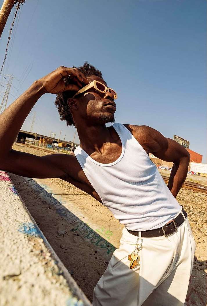 Black man wearing a white tank top, cream sunglasses, and white trousers leaning outdoors under clear sky.