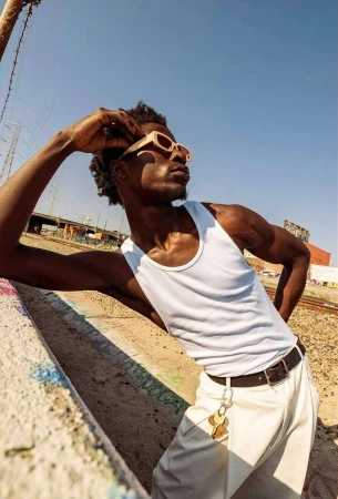 Black man wearing a white tank top, cream sunglasses, and white trousers leaning outdoors under clear sky.