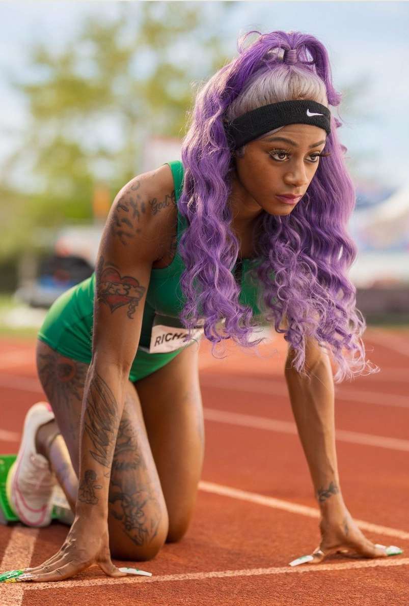 An adult female athlete with purple hair is on a running track in a three-point start position, preparing to race, wearing a green athletic uniform.