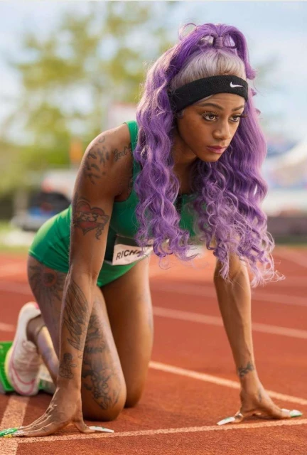 An adult female athlete with purple hair is on a running track in a three-point start position, preparing to race, wearing a green athletic uniform.