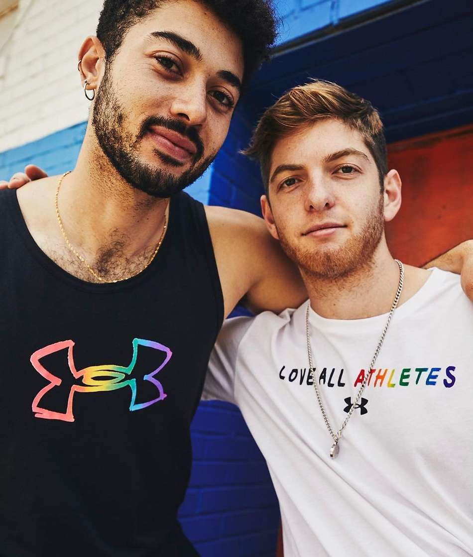 Two adult males pose indoors, wearing Under Armour branded apparel with rainbow logo detail, medium shot, eye-level view, good lighting.