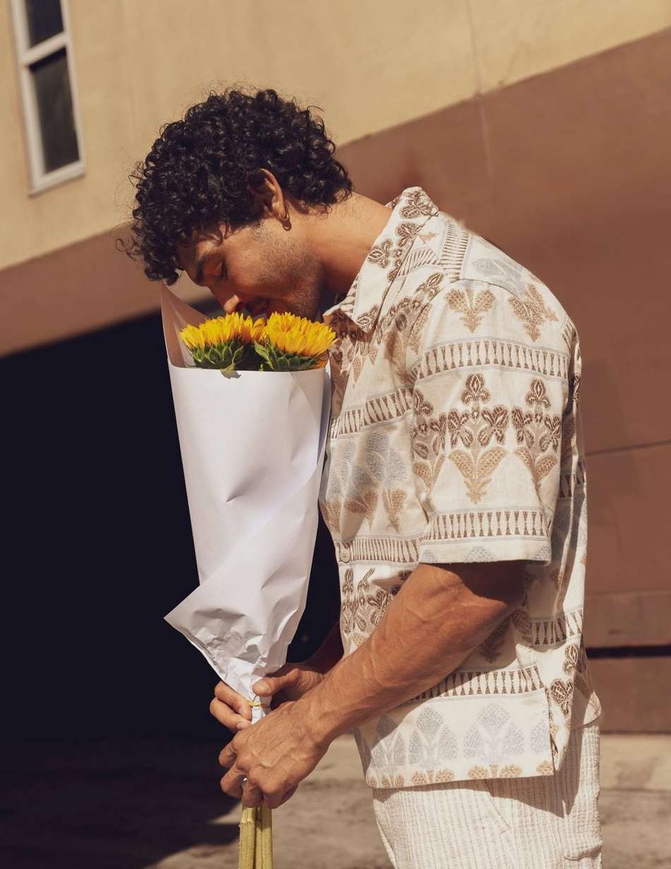 Curly-haired man in patterned shirt smells a bouquet of bright yellow sunflowers in a white paper wrap, outside on a sunny day.