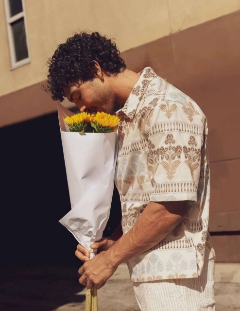 Curly-haired man in patterned shirt smells a bouquet of bright yellow sunflowers in a white paper wrap, outside on a sunny day.