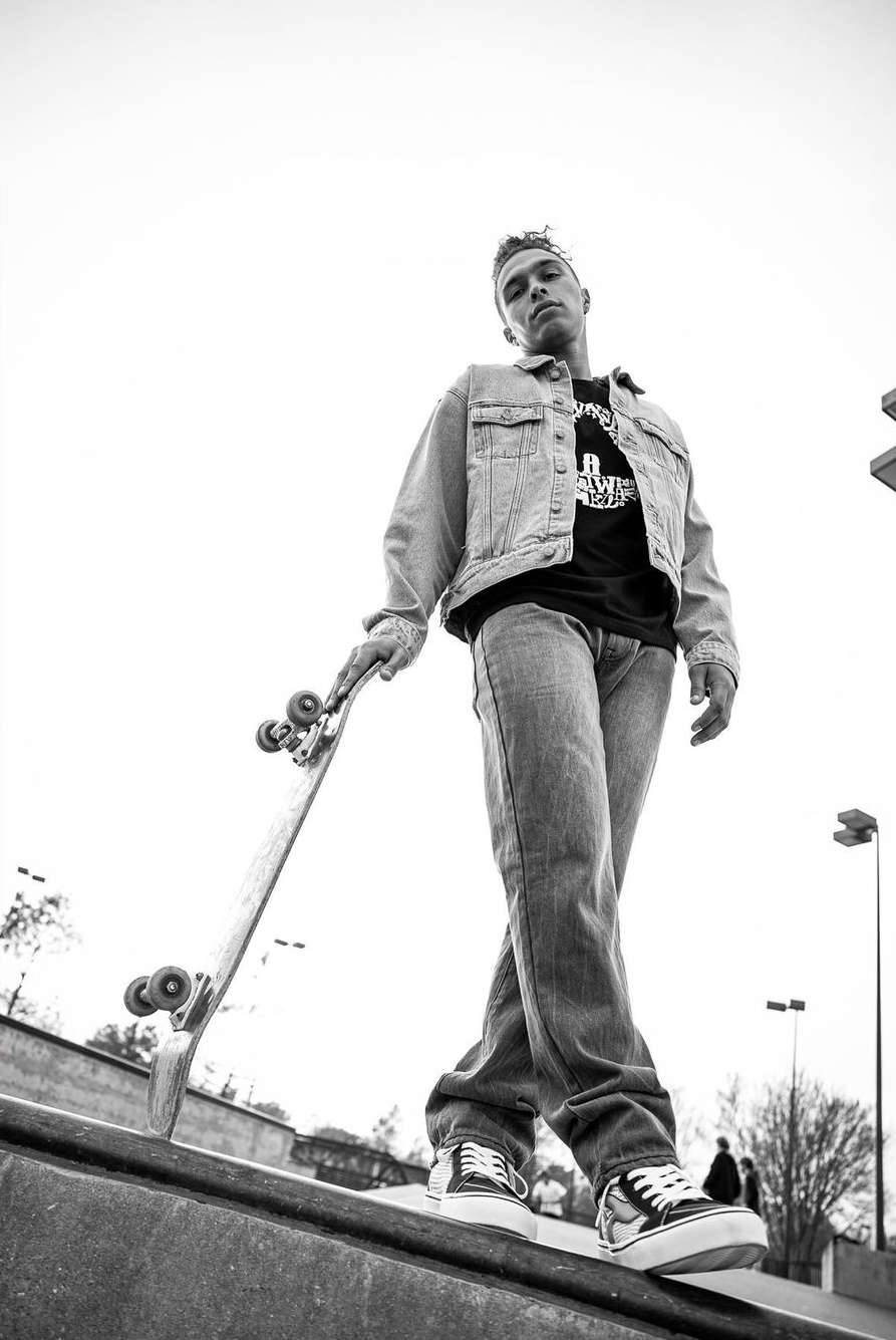 Black and white shot of a young adult male in jeans and a denim jacket, holding a skateboard and standing in a skate park.