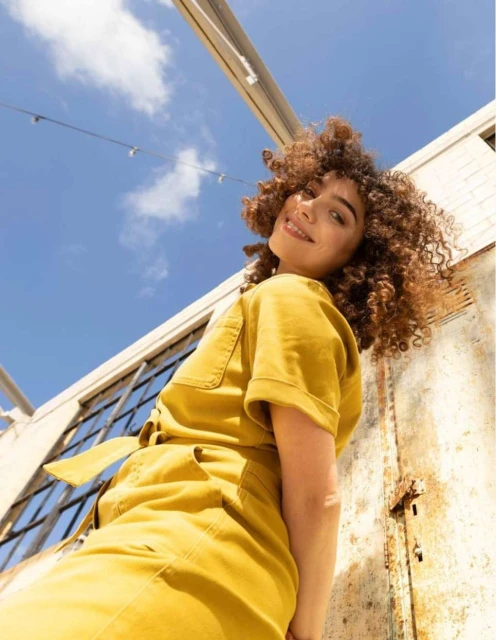A young woman with curly hair smiles at the camera, wearing a yellow jumpsuit outdoors with a partially cloudy sky above her.
