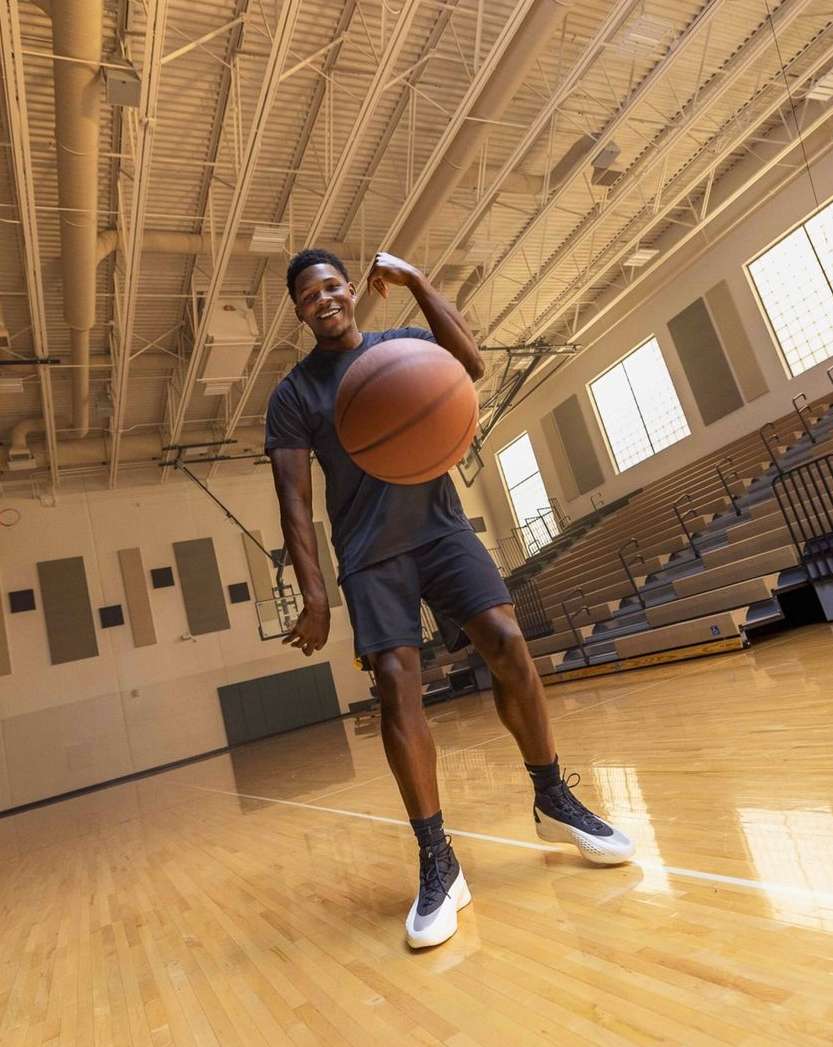 An African American male in athletic wear bounces a basketball in an indoor gymnasium with hardwood floors and overhead lighting.