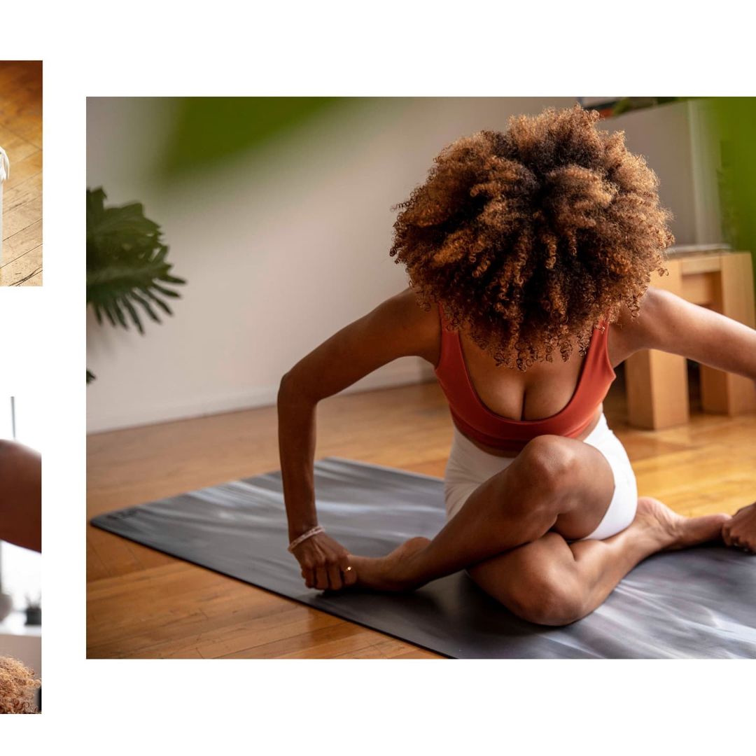 Woman with curly hair wearing a sports bra and shorts doing yoga on a mat in a home interior with wood floors and natural lighting.