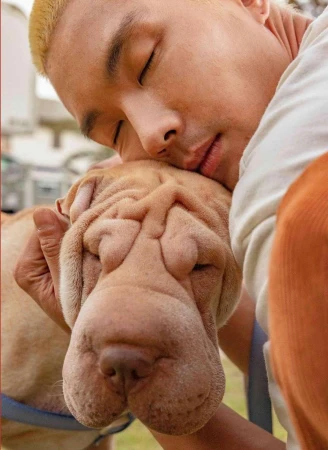 Close-up portrait of a fair-skinned man with blond hair embracing a Shar Pei dog with a wrinkled face, both with their eyes closed.