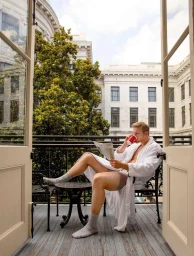 Man in white bathrobe reading a newspaper and drinking from a red cup on a hotel balcony in New York